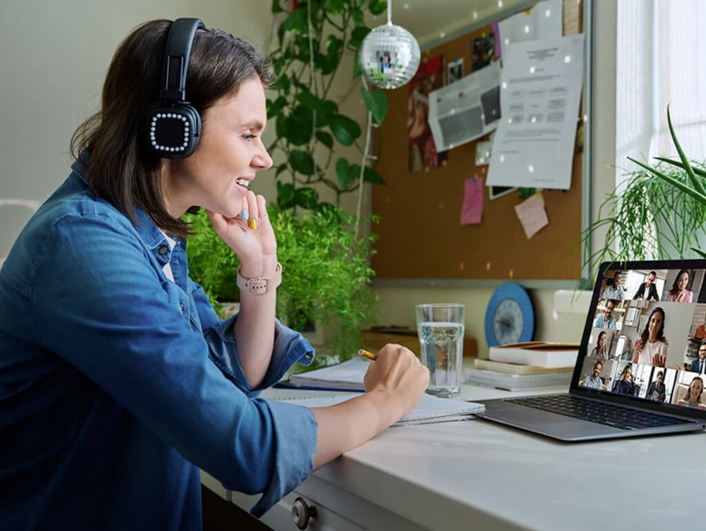 Woman wearing headphones, smiling, and taking notes during a video conference on a laptop at a home office desk.