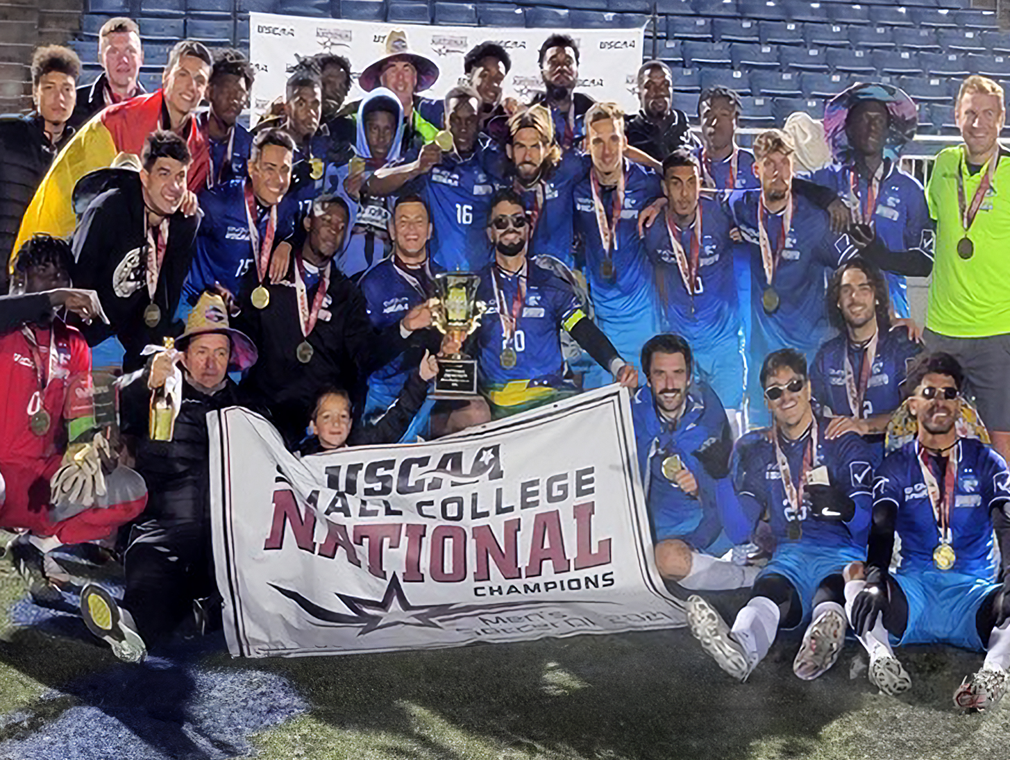 A college soccer team celebrates winning the national championship, holding medals, a trophy, and a championship banner.