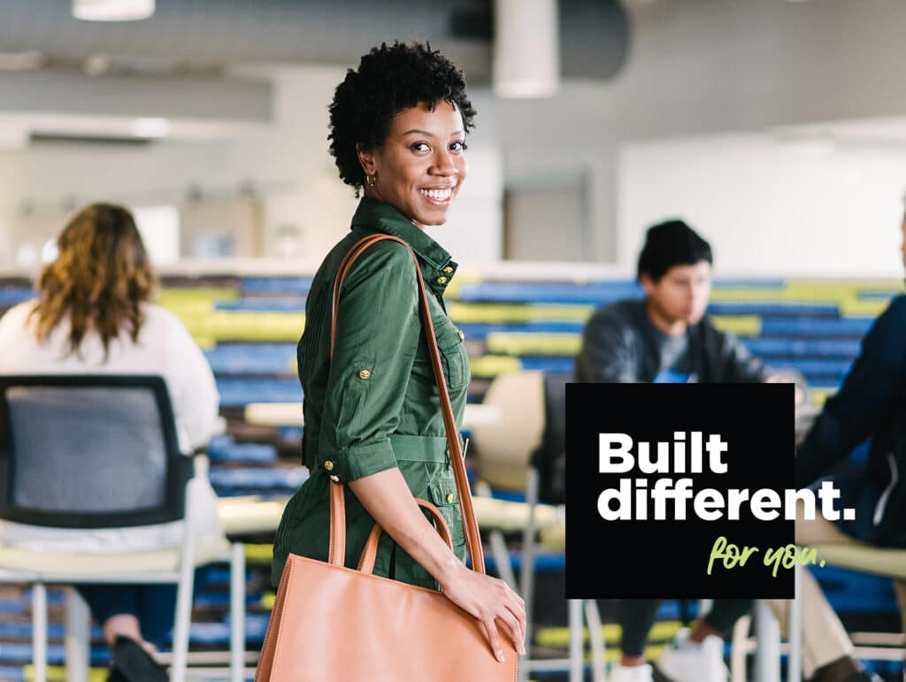 Smiling person in a green jacket carrying a brown bag, with "Built different. for you." text overlay, in a casual work setting.