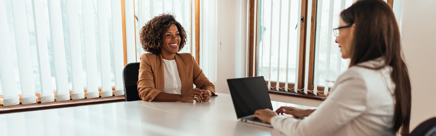 Two women having a conversation at a table, one typing on a laptop and the other smiling, in a bright office setting.