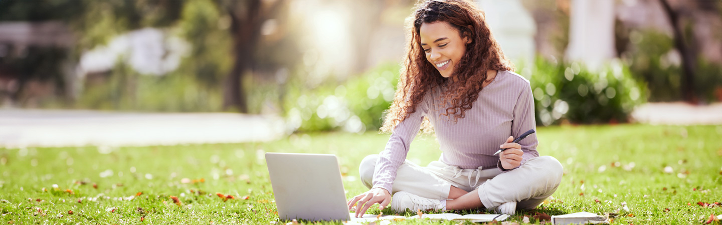 Woman sitting cross-legged on grass, working on a laptop, smiling, with notebooks and pen in hand on a sunny day.