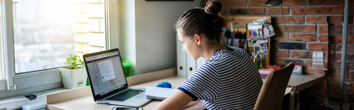 Person with a bun hairstyle working on a laptop at a desk with a notebook, plants, and office supplies in a well-lit room.