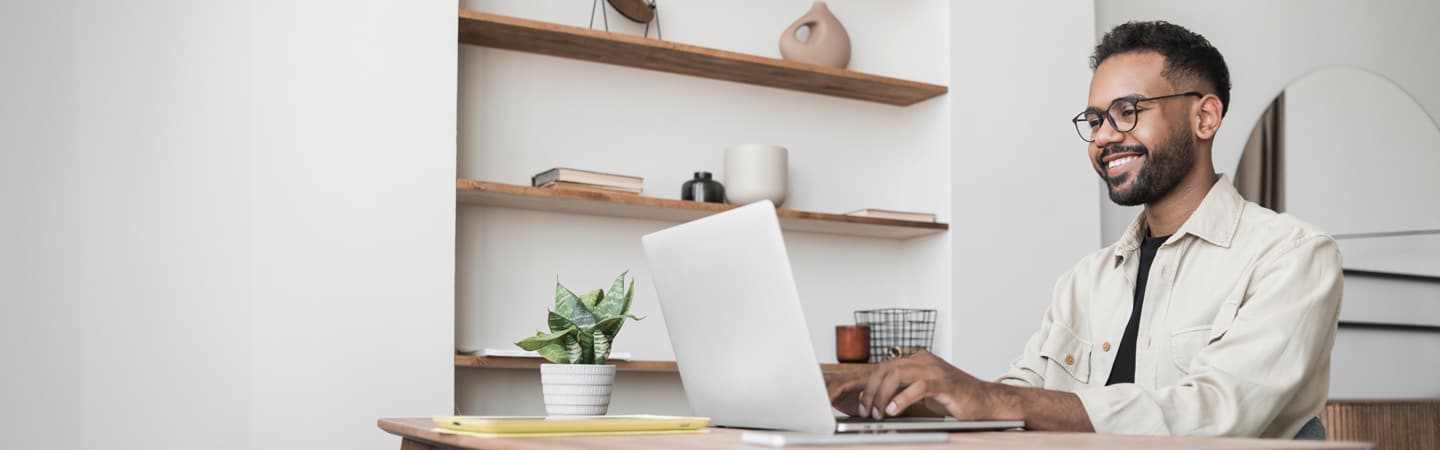 A man sits at a desk, smiling while using a laptop. A plant and a notebook are on the desk. Shelves are in the background.
