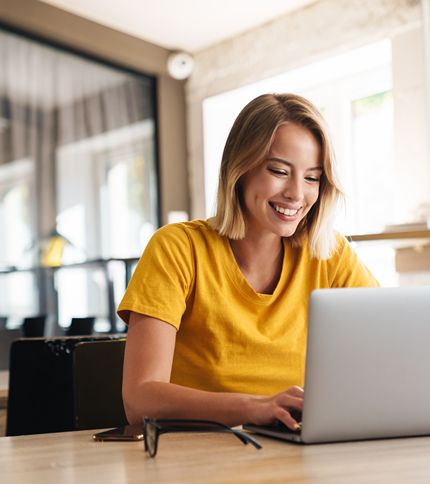 A young Social Media Coordinator in a yellow shirt is smiling while working on a laptop at a wooden table in a bright, modern room. Glasses rest on the table beside her.