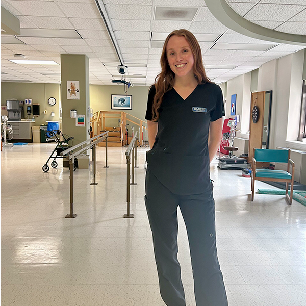 A person stands smiling in a medical uniform in a brightly lit, clean hospital or clinic setting.