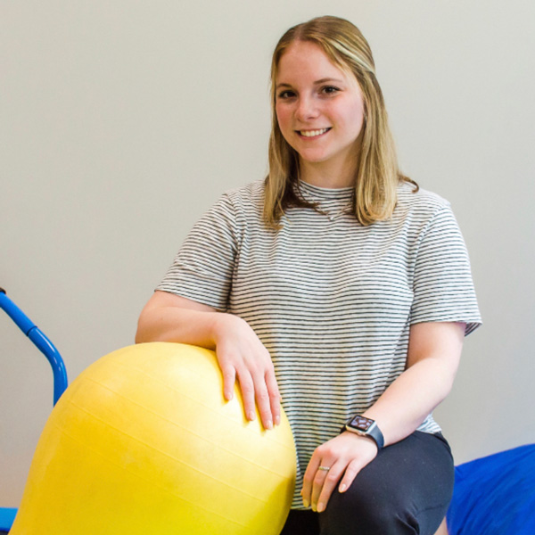 A woman with blonde hair, wearing a striped shirt, smiles while resting her hand on a yellow exercise ball.