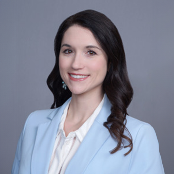 Smiling woman with long dark hair wearing a light blue blazer and white shirt against a gray background.