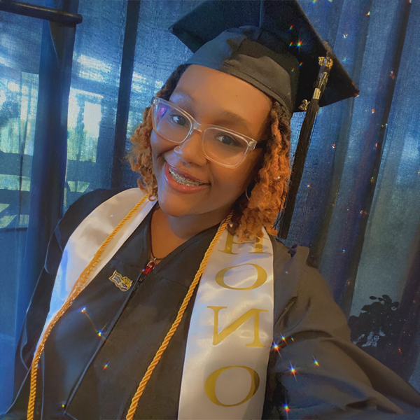 A graduate wearing a cap and gown with a white honor stole smiles in front of a blue curtain.