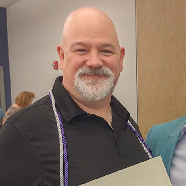 A man with a bald head and white beard, wearing a black shirt and a cord, smiles while holding a certificate.