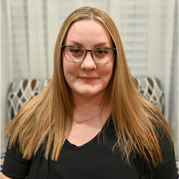 A woman with long straight blonde hair, glasses, and a black shirt, standing in front of a patterned curtain.