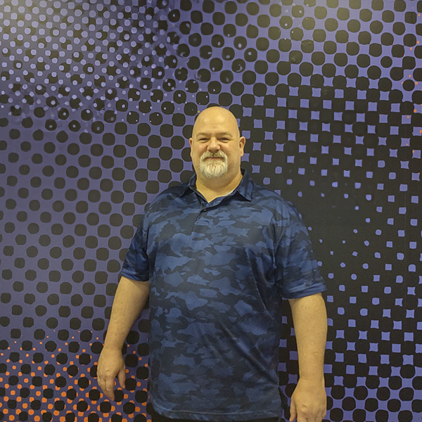A man with a beard and mustache stands in front of a purple and black dotted background, wearing a blue camo shirt.