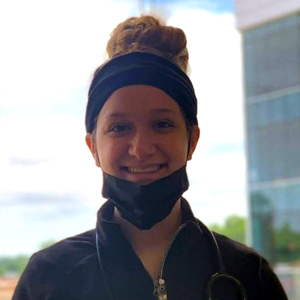 Smiling person with a bun hairstyle and headband, standing outdoors in front of a building.