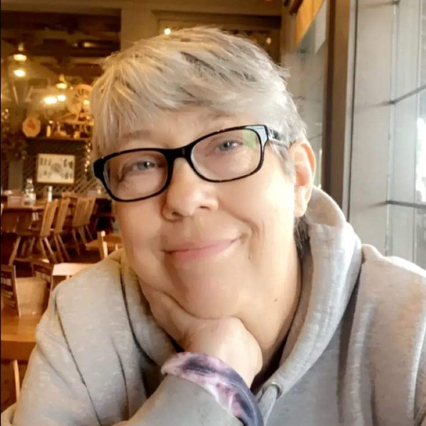 A person with gray hair and glasses smiles, resting their chin on their hand, seated in a cozy, warmly-lit room.