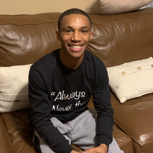 Smiling person in a black shirt with text sits on a brown leather couch, with white pillows behind them.