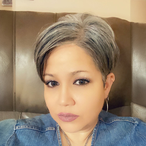 A woman with short, gray hair and hoop earrings, wearing a denim shirt, sits on a brown leather couch.