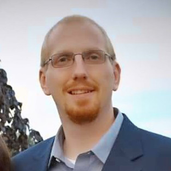 A smiling man with short light hair, glasses, and a goatee, dressed in a suit jacket, against an outdoor background.