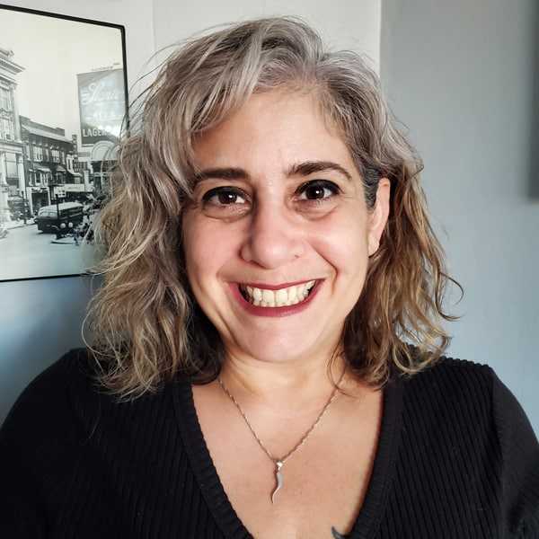 Smiling woman with wavy gray hair and a black sweater poses in front of a vintage photograph background.