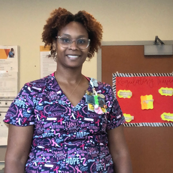A person in colorful scrubs stands smiling in a room with bulletin boards in the background.