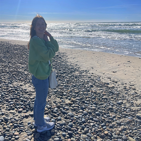A person in a green jacket stands on a rocky beach looking at the ocean under a clear, sunny sky.