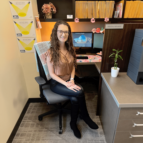 Woman seated at an office desk with a computer, files, and decor, smiling at the camera.