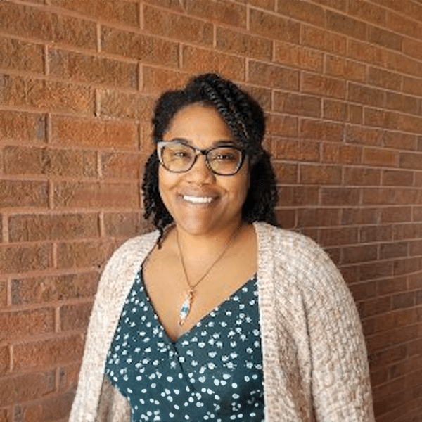 Person with glasses and curly hair smiling, wearing a patterned dress and beige sweater against a brick wall background.