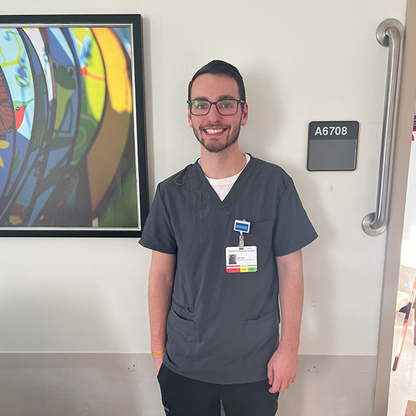 Smiling person in scrubs with ID badge stands by a wall sign labeled "A6708," colorful artwork in the background.