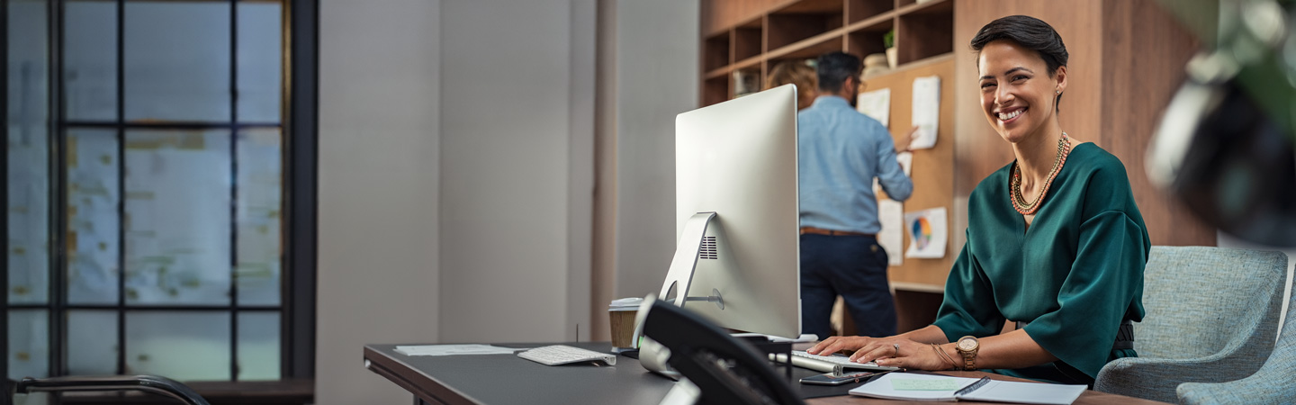 A woman smiling at her desk with a computer, while a man works on a bulletin board in the background.