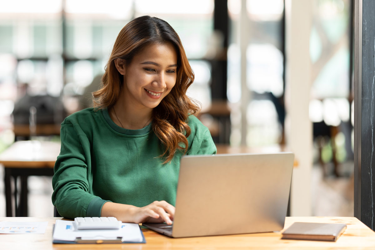 Woman in a green sweater smiling while typing on a laptop at a desk in a bright, modern workspace.