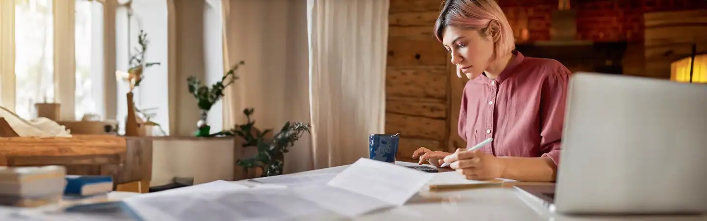 Person with pink hair working on a laptop at a desk with papers and a mug, in a cozy, well-lit room.