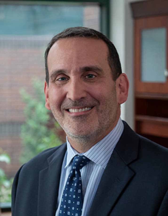 A smiling man in a suit and tie stands in an office with a bookshelf and a window in the background.