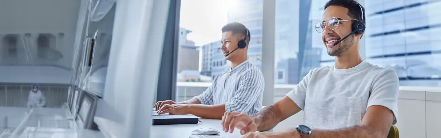 Two men wearing headsets and working on computers in a bright office environment with large windows.