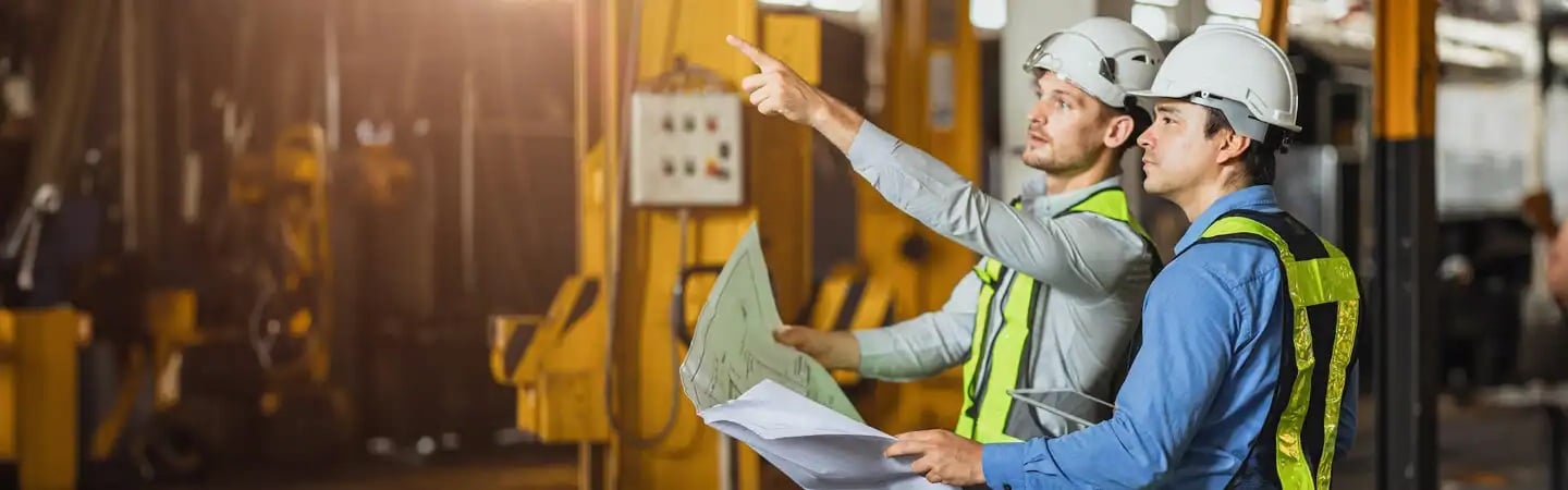 Two construction workers in safety vests and helmets examine blueprints and point in a warehouse or industrial setting.