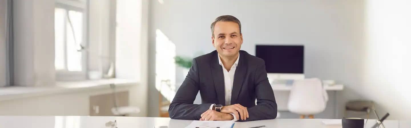 A smiling man in a suit sits at a bright, modern office desk with a computer in the background.
