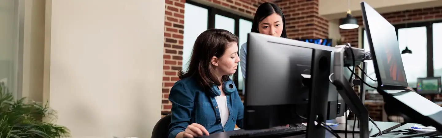 Two women working at a computer desk in a modern office with brick walls and large windows.