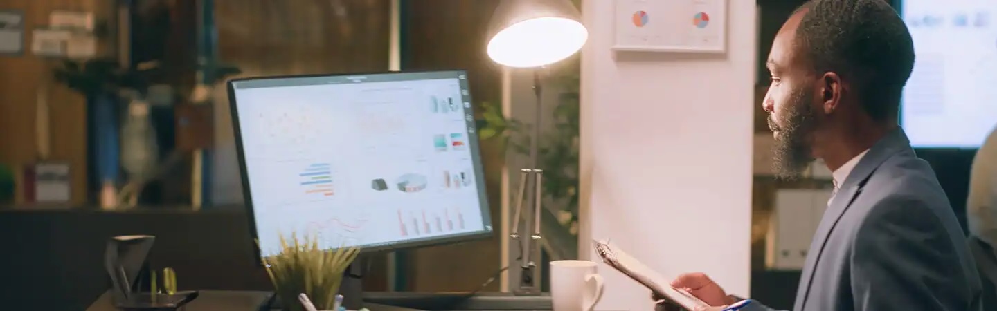 A man in a suit analyzes charts on a computer screen, holding a clipboard, in a modern office.