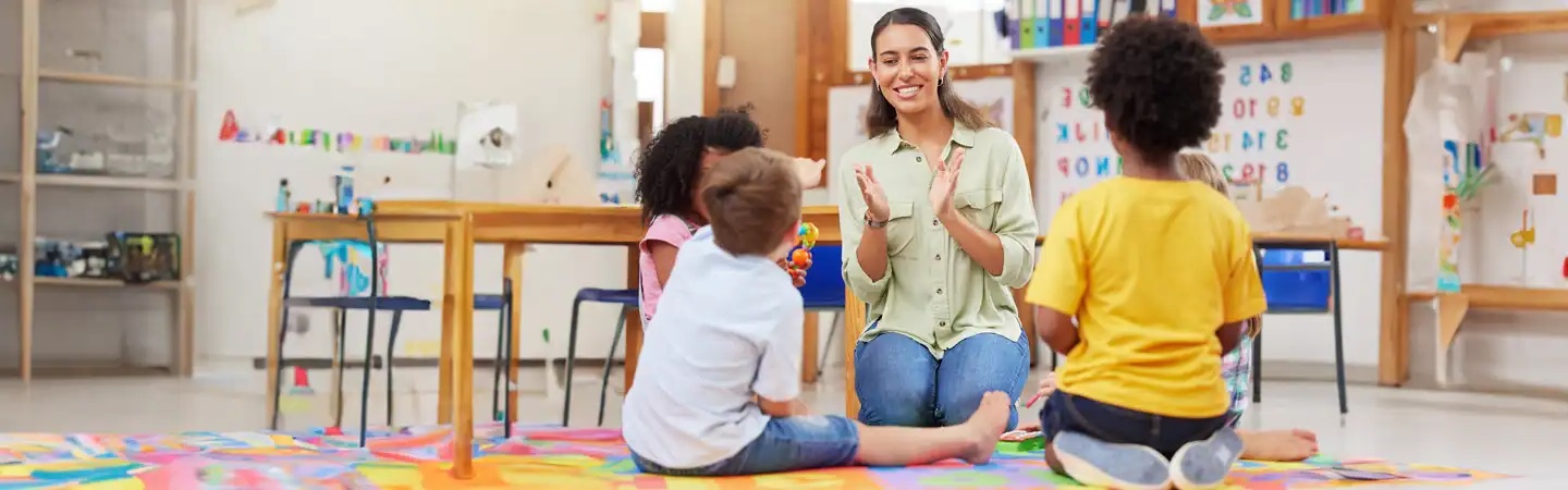 A teacher claps and sits on the floor with three young children in a colorful classroom.