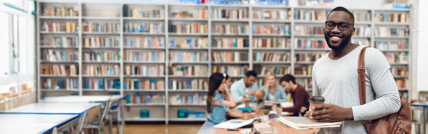 Man with coffee cup and books smiling in a library, with others studying in the background.