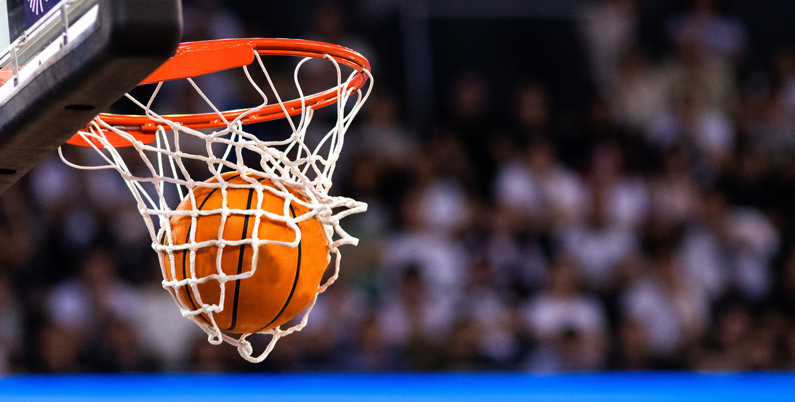 Close-up of a basketball going through a hoop with a net, with a crowd of spectators blurred in the background.