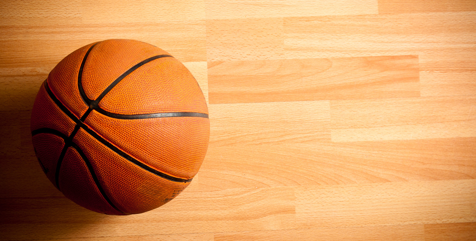 A basketball resting on a hardwood court, illuminated from the side.
