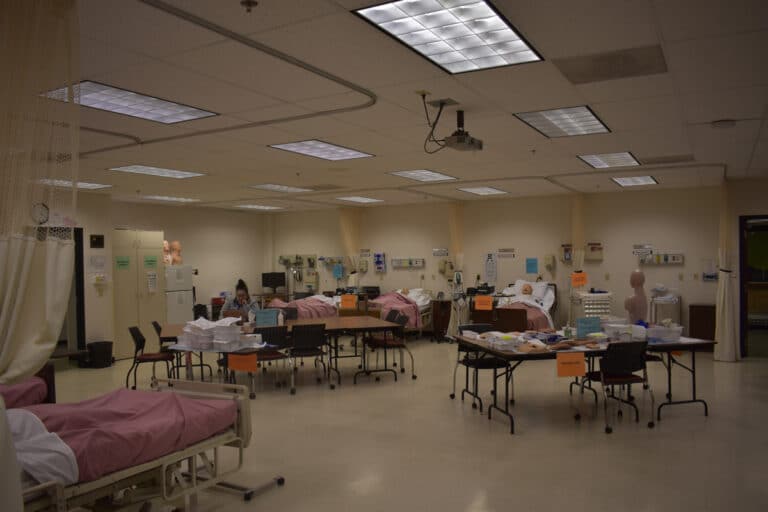 Large, brightly-lit medical training room with hospital beds, mannequins, and tables covered in medical supplies.