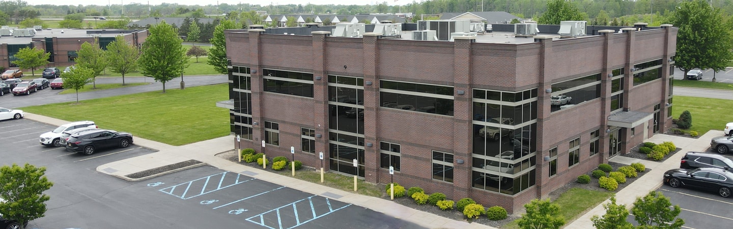 Aerial view of a two-story brick office building with parking spaces and a few cars around it, surrounded by greenery.