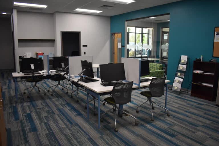 A modern office space with desks, computers, chairs, and a bookshelf against a blue and gray wall.