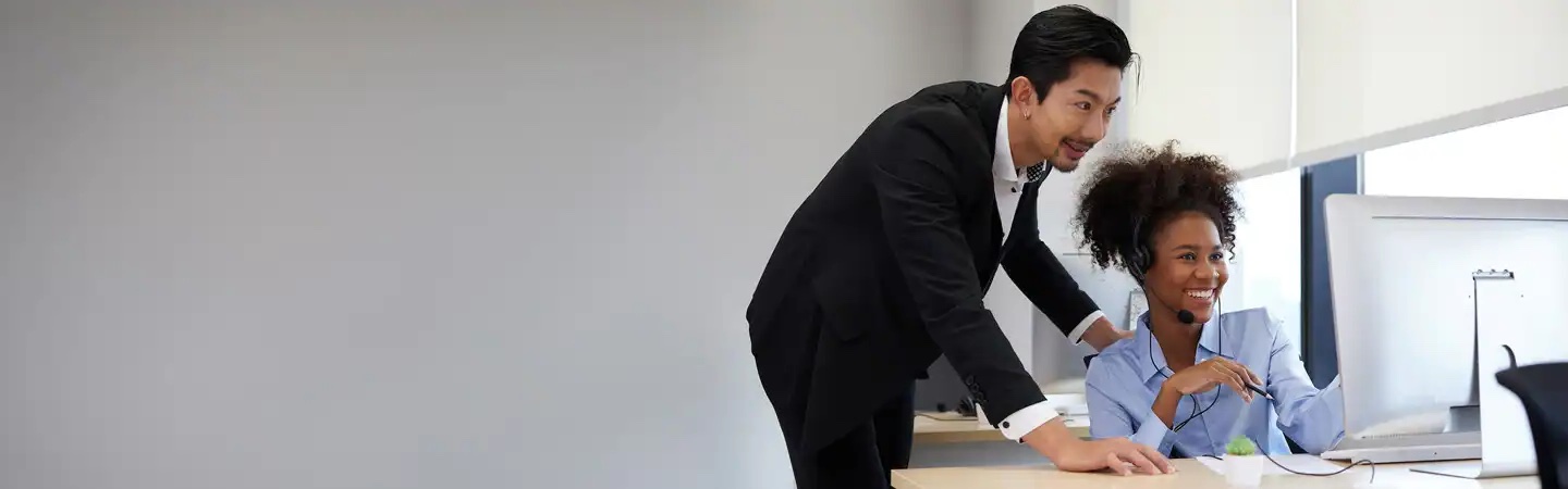 A man in a suit leans over to assist a woman wearing a headset, smiling, in a modern office setting.