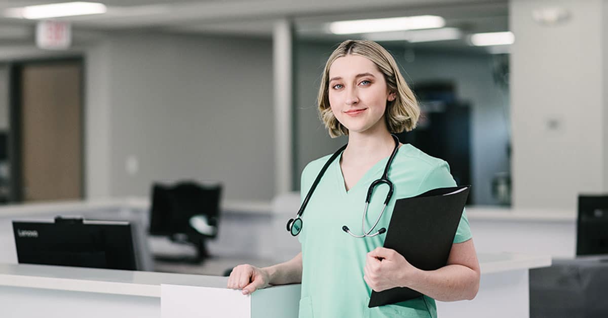 A nurse in green scrubs stands confidently at a hospital desk, holding a clipboard with a stethoscope around her neck.
