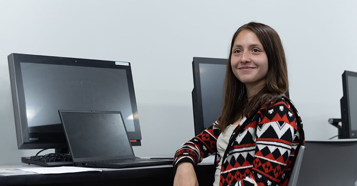 A person sits at a desk with a laptop, smiling at the camera, in a room with desktop computers in the background.