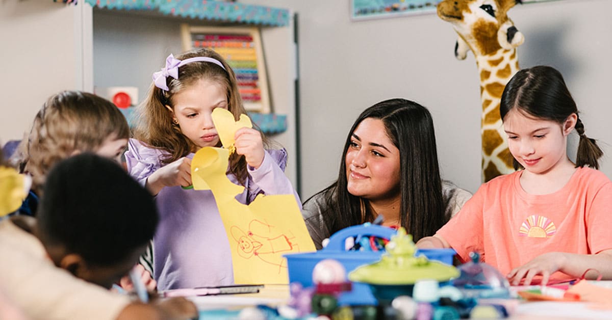 Children and an adult at a table engaged in arts and crafts, with a large toy giraffe in the background.