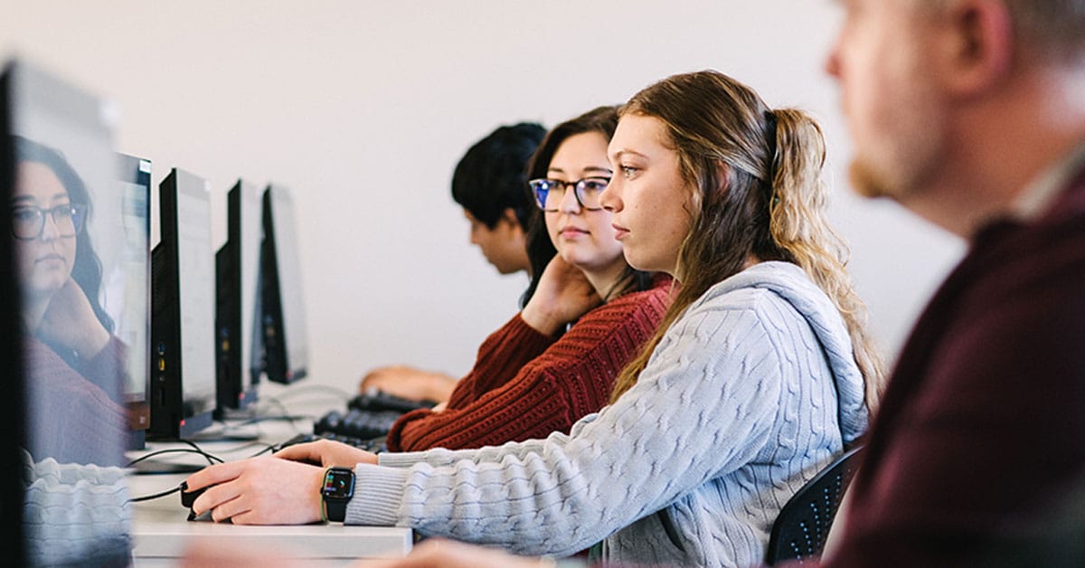 People sitting in a row at computer desks, focused on their screens, in a classroom or office setting.