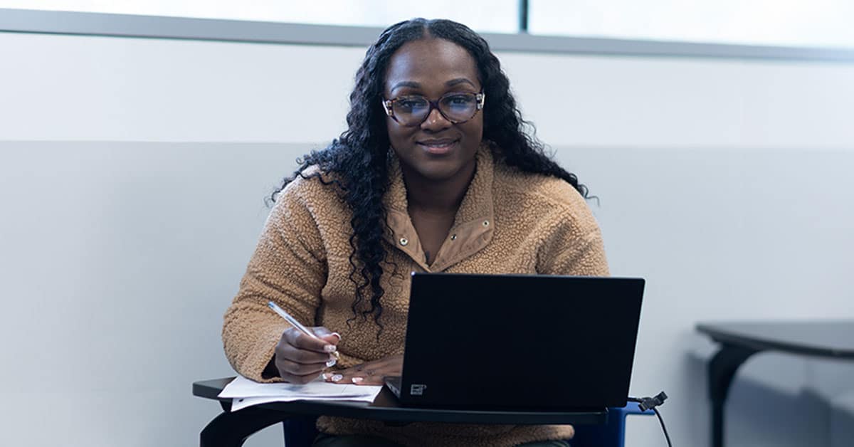 A person sitting at a desk with a laptop, writing in a notebook and smiling.