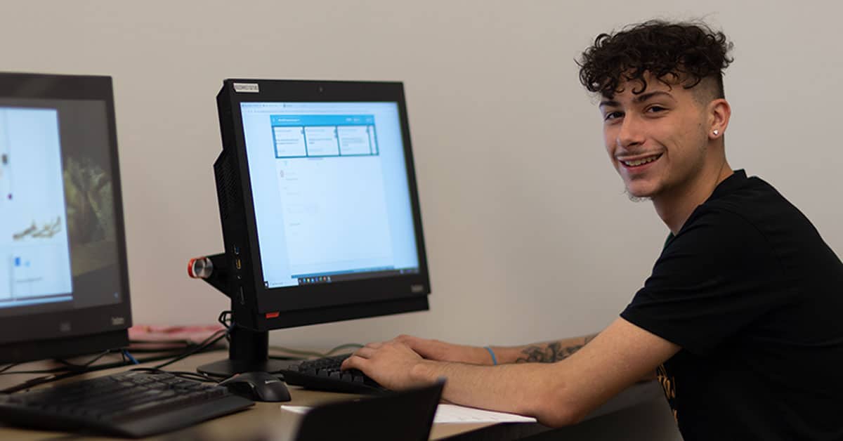 A person with curly hair sits at a desk, smiling, and working on a computer in an office or classroom setting.
