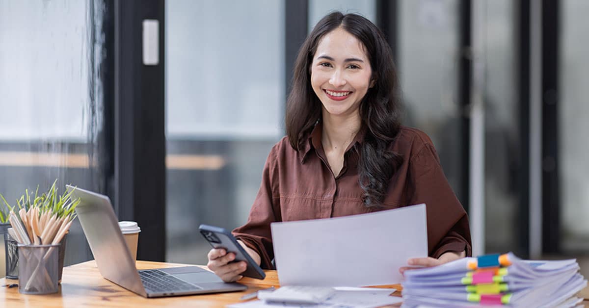 A person sitting at a desk with a laptop, holding a document and a phone, smiling, and working.